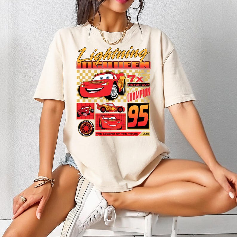 May include: Beige t-shirt with a graphic of Lightning McQueen from the Cars film. The design includes the text "Lightning McQueen" and "7x Piston Cup Champion 95". The shirt has short sleeves and a crew neck.