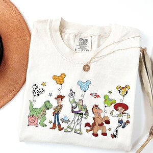 May include: Cream-colored t-shirt featuring a colorful graphic of animated characters, including a dinosaur, pig, cowboy, spaceman, and cowgirl. The design includes balloons and stars. The shirt has a crew neck and short sleeves.