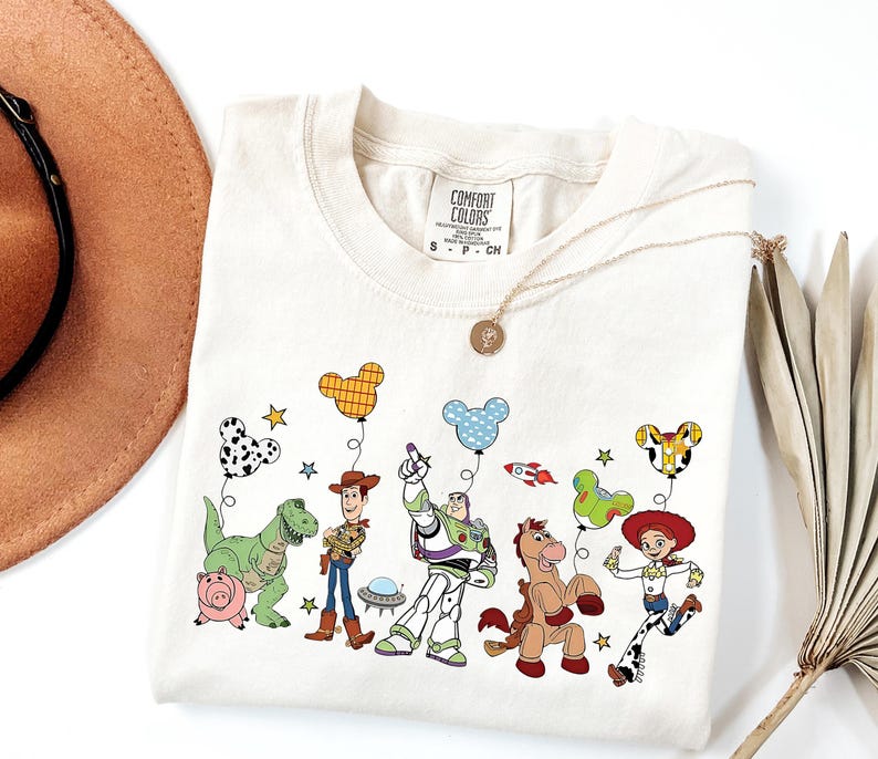 May include: Cream-colored t-shirt featuring a colorful graphic of animated characters, including a dinosaur, pig, cowboy, spaceman, and cowgirl. The design includes balloons and stars. The shirt has a crew neck and short sleeves.