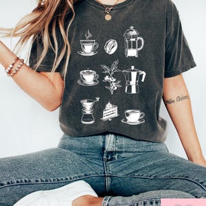 May include: Dark grey t-shirt with white coffee-themed illustrations, such as cups, beans, and a cafeti&egrave;re. The shirt also has the text "Magdalene Giftshopco Est 2024" at the bottom.