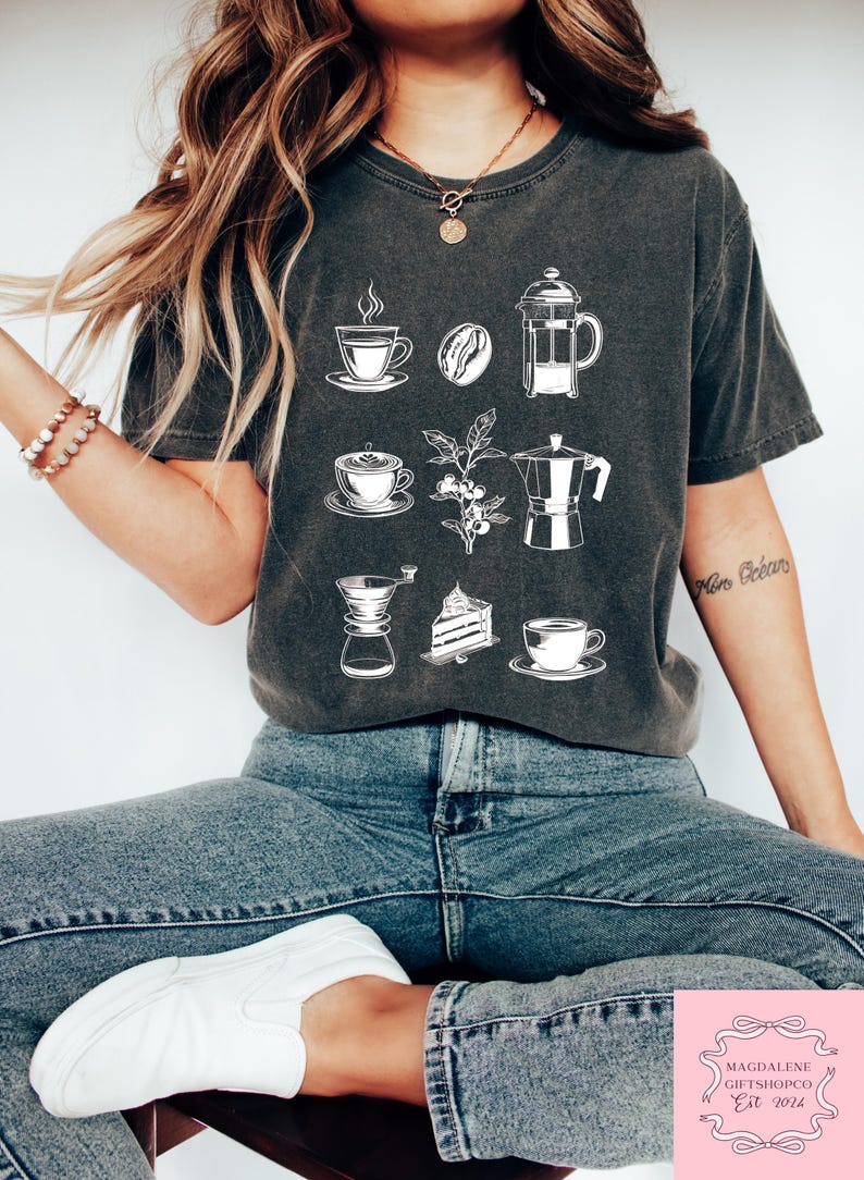 May include: Dark grey t-shirt with white coffee-themed illustrations, such as cups, beans, and a cafeti&egrave;re. The shirt also has the text "Magdalene Giftshopco Est 2024" at the bottom.