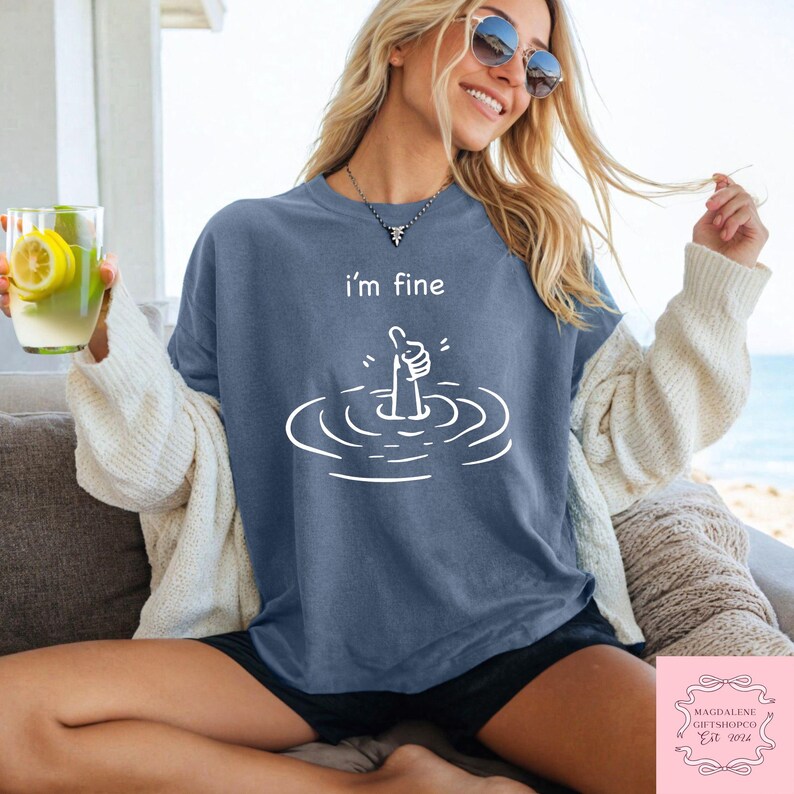 May include: A blue t-shirt with a white graphic of a hand giving a thumbs up while submerged in water. The text "i'm fine" is printed above the graphic.