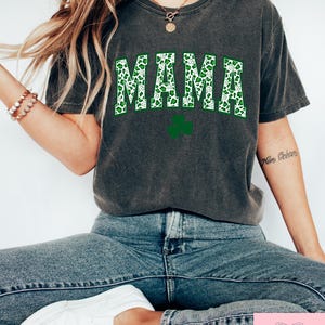 May include: A dark gray t-shirt with the word "MAMA" printed in white letters with a green shamrock pattern. The shamrock pattern is a green and white leopard print.