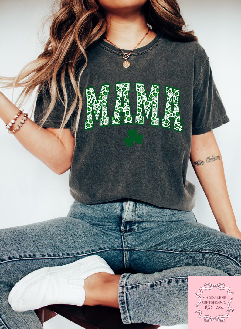 May include: A dark gray t-shirt with the word "MAMA" printed in white letters with a green shamrock pattern. The shamrock pattern is a green and white leopard print.
