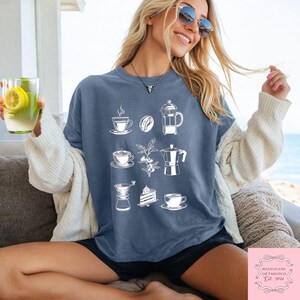 May include: A blue t-shirt with white illustrations of coffee-related items, such as cups, beans, a French press, and a moka pot. The shirt is worn by a person with sunglasses and a white cardigan. The text "MAGDALENE GIFTSHOPCO Est. 2024" is in the bottom right corner.