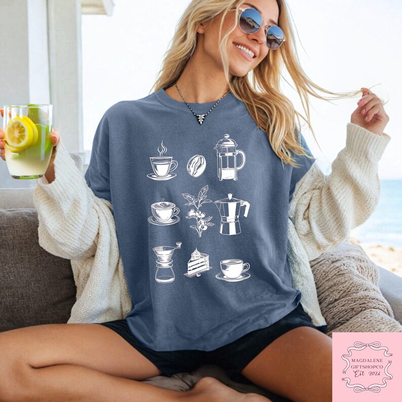 May include: A blue t-shirt with white illustrations of coffee-related items, such as cups, beans, a French press, and a moka pot. The shirt is worn by a person with sunglasses and a white cardigan. The text "MAGDALENE GIFTSHOPCO Est. 2024" is in the bottom right corner.
