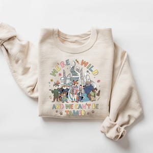 May include: A cream-colored sweatshirt with the text "WE'RE WILD AND WE CAN'T BE TAMED" in colorful letters. The graphic features a group of cartoon animals and a cityscape design. The sweatshirt has long sleeves.