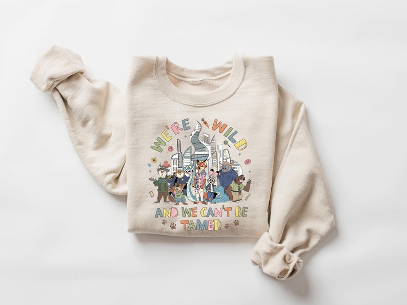May include: A cream-colored sweatshirt with the text "WE'RE WILD AND WE CAN'T BE TAMED" in colorful letters. The graphic features a group of cartoon animals and a cityscape design. The sweatshirt has long sleeves.