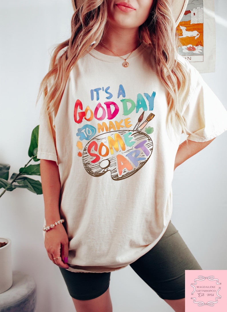May include: Cream-colored t-shirt with the text "It's a Good Day to Make Some Art" in colorful letters. The design includes a painter's palette with paintbrushes. The shirt is a casual, artistic style.