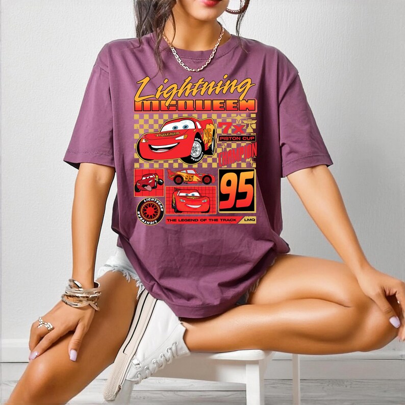 May include: A purple t-shirt with a graphic of Lightning McQueen from the film Cars. The design includes the text "Lightning McQueen" in yellow, the number "95", and images of the character. The shirt is short-sleeved and casual.