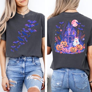 May include: A charcoal gray short-sleeve t-shirt featuring a front graphic of blue and purple bats in flight. The back graphic displays a purple haunted house, a white ghost with a pink bow, orange jack-o'-lanterns, and a pink moon.