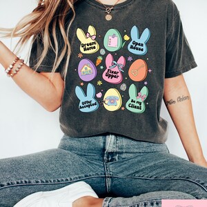 May include: A dark gray t-shirt with a colorful Easter egg design. The eggs feature phrases like "Dream Home", "Open House", "Fixer Upper", "Offer Accepted", and "Be My Client".