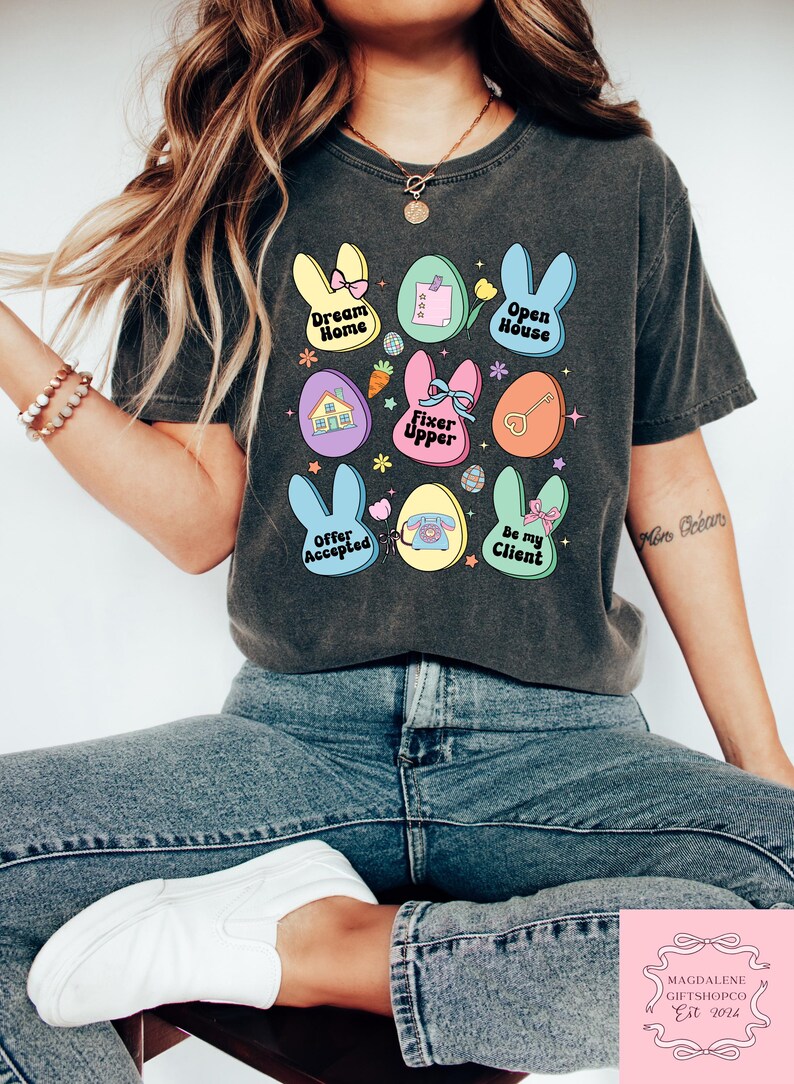 May include: A dark gray t-shirt with a colorful Easter egg design. The eggs feature phrases like "Dream Home", "Open House", "Fixer Upper", "Offer Accepted", and "Be My Client".