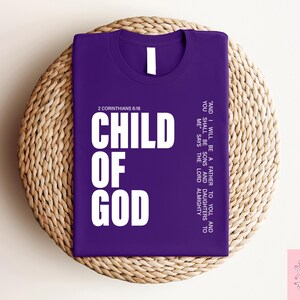 Peut inclure: Un t-shirt violet avec l'inscription "CHILD OF GOD" en grandes lettres blanches. Le texte "2 Corinthiens 6:18" est au-dessus des mots. Le t-shirt affiche &eacute;galement une citation. Le t-shirt est pos&eacute; sur un tapis rond en osier.