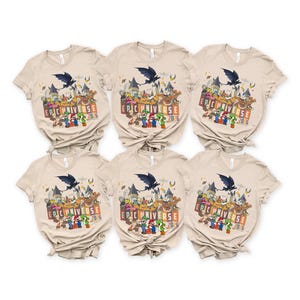 May include: Six beige short-sleeved t-shirts with a colorful graphic design. The design includes a castle, cartoon characters, a bat, and the text "EPIC UNIVERSE 2026". The shirts are neatly arranged.
