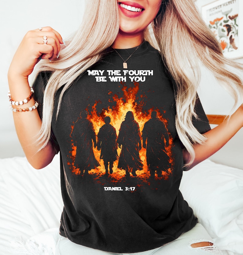 K&ouml;nnte beinhalten: Schwarzes T-Shirt mit wei&szlig;em Text "MAY THE FOURTH BE WITH YOU". Die Grafik zeigt vier Figuren vor einem feurigen Hintergrund, darunter "DANIEL 3:17". Ein Star Wars-Design.