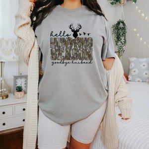 K&ouml;nnte beinhalten: Graues T-Shirt mit dem Text "hello HUNTING SEASON goodbye husband" in Schwarz und Camouflage-Print. Dar&uuml;ber befinden sich eine schwarze Hirschsilhouette und V&ouml;gel. Das Shirt wird mit wei&szlig;en gerippten Shorts und einer cremefarbenen Strickjacke getragen.