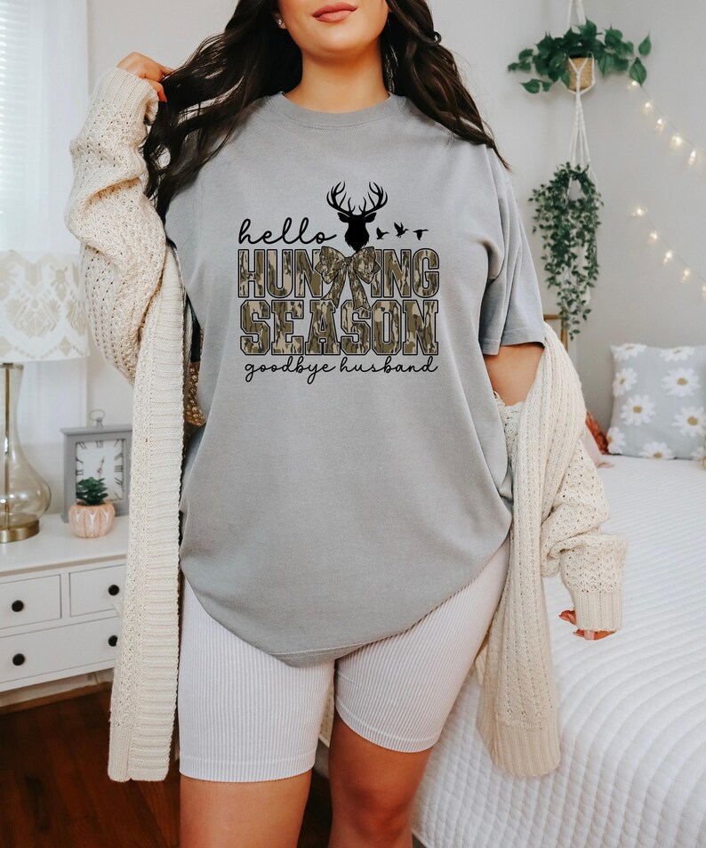 K&ouml;nnte beinhalten: Graues T-Shirt mit dem Text "hello HUNTING SEASON goodbye husband" in Schwarz und Camouflage-Print. Dar&uuml;ber befinden sich eine schwarze Hirschsilhouette und V&ouml;gel. Das Shirt wird mit wei&szlig;en gerippten Shorts und einer cremefarbenen Strickjacke getragen.