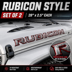 以下が含まれることがあります： 車両のボンネット用Rubiconデカール2枚セット。各サイズは71.12cm x 6.35cmです。デカールは金属のような質感で、赤い輪郭線が施されています。テキストはRubiconと書かれています。円形のインセットには文字Rのクローズアップが表示されています。
