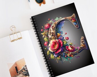 Pode incluir: Um caderno espiral preto com um design floral colorido que envolve uma letra C dourada adornada. O fundo é um degradé cinza escuro.