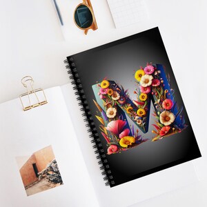 May include: A spiral notebook with a black cover featuring a colorful floral letter M. The letter is made up of various flowers and leaves in shades of pink, yellow, blue, and green.