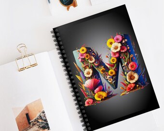 Pode incluir: Um caderno espiral com capa preta apresentando uma letra M floral colorida. A letra é composta por várias flores e folhas em tons de rosa, amarelo, azul e verde.