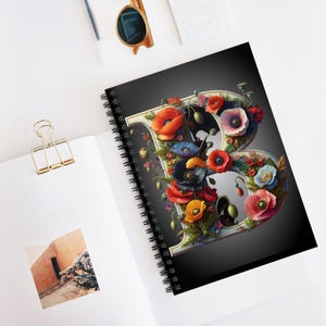 May include: A spiral bound notebook with a black cover featuring a large letter "B" made of white paper with a floral design. The flowers are red, orange, yellow, pink, and blue.