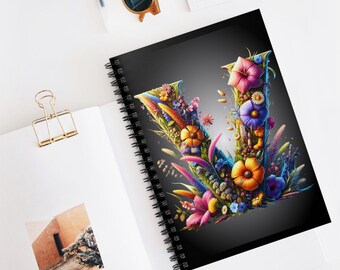 May include: A spiral-bound notebook with a black cover featuring a floral letter "V". The letter is adorned with colorful flowers in shades of pink, orange, yellow, and blue. The notebook is open on a white surface.