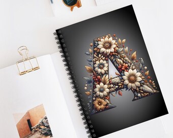 May include: A spiral bound notebook with a black cover featuring a large letter A made of colorful flowers. The letter A is surrounded by a dark gray background.