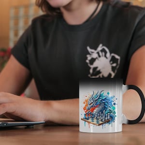 May include: A white ceramic mug with a colourful dragon design playing chess. The dragon is depicted with blue, orange, and red hues. The mug has a black handle and a black interior. The mug is on a wooden table.