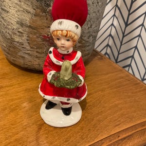 May include: A vintage ceramic figurine of a child in a red and white Santa Claus outfit, holding a green wreath. The figurine has a red velvet pin cushion hat. The base is white, and the figurine is on a wooden surface.