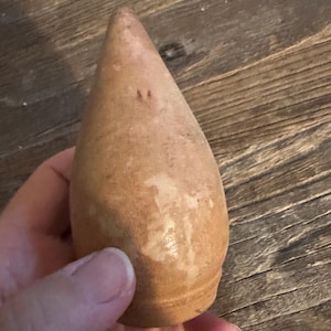 May include: A light brown, teardrop-shaped wooden object with a smooth, tapered point at the top and a rounded base. The surface shows natural wood grain variations and light-colored patches, with a small indentation near the apex.