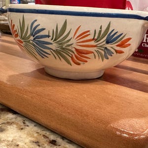 May include: A ceramic bowl with a white base, adorned with a floral pattern in orange, blue, and green. The bowl has a blue rim and handles. It sits on a wooden cutting board.