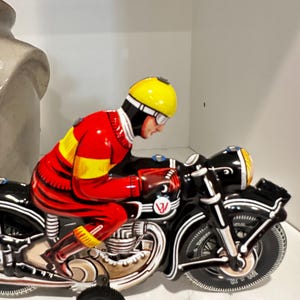 Könnte beinhalten: Ein Spielzeugmotorrad aus Blech im Vintage-Stil mit einem Fahrer in rotem und gelbem Rennanzug und einem leuchtend gelben Helm. Das Motorrad ist schwarz mit weißen Zierlinien, einem runden Logo mit einem roten W und einem kleinen schwarzen Stützrad.