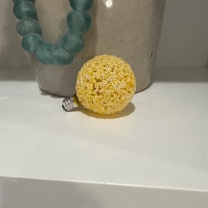 May include: A yellow, textured decorative light bulb with a silver base rests on a white surface. A strand of blue-green glass beads hangs near a beige ceramic jug.