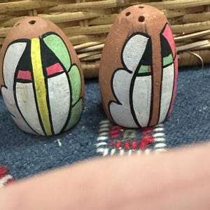 May include: Two brown, egg-shaped salt and pepper shakers. Each shaker has a painted design featuring white, green, yellow, black, and pink shapes. The shakers have three holes on top for dispensing.