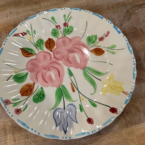 May include: A white ceramic plate with a scalloped edge and a hand-painted floral design. The plate features pink flowers, green leaves, and accents of yellow, blue, and red. The rim is decorated with a blue dotted border.