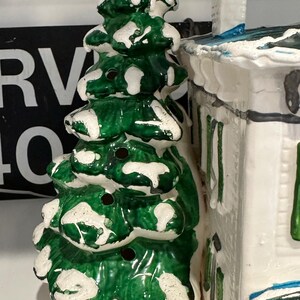 K&ouml;nnte beinhalten: Eine Keramik-Weihnachtsbaumfigur mit gr&uuml;ner und wei&szlig;er Glasur. Der Baum hat einen schneebedeckten Sockel und ist mit wei&szlig;en Punkten verziert. Die Figur steht neben einem wei&szlig;en Keramikhaus mit blauem Dach.