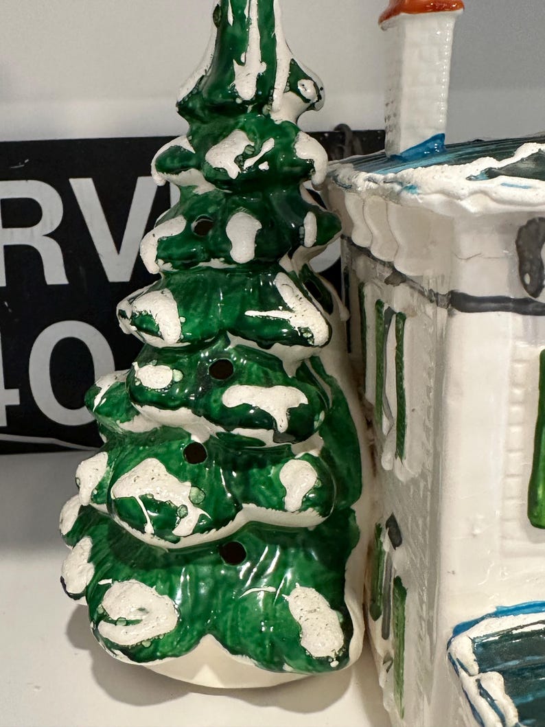 K&ouml;nnte beinhalten: Eine Keramik-Weihnachtsbaumfigur mit gr&uuml;ner und wei&szlig;er Glasur. Der Baum hat einen schneebedeckten Sockel und ist mit wei&szlig;en Punkten verziert. Die Figur steht neben einem wei&szlig;en Keramikhaus mit blauem Dach.
