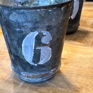 Może przedstawiać: Dwa zwietrzałe metalowe wiadra z postarzanym wykończeniem. Każde wiadro ma białą, namalowaną literę. Wiadra stoją na drewnianej powierzchni.