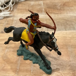 Peut inclure: Figurine en plastique d'un guerrier amérindien à cheval, tirant à l'arc. Le guerrier porte une tenue bleue et jaune, et le cheval est noir avec une face blanche. La base est verte.
