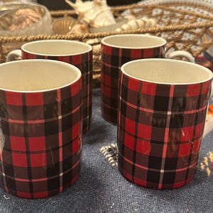 Puede incluir: Cuatro tazas cilíndricas con un patrón de cuadros rojos y negros. Las tazas tienen interiores blancos y asas pequeñas. Están dispuestas sobre una superficie azul estampada, lo que sugiere un tema rústico y acogedor.