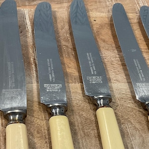 May include: Five silver table knives with ivory-colored handles. The knives are engraved with the text "First Sheffield Stainless" and "Made in China" on the blades.