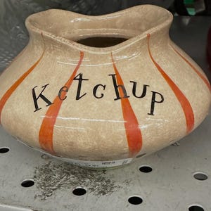 May include: A beige ceramic ketchup dispenser with orange vertical stripes and the word "Ketchup" in black lettering. The dispenser has a wide opening and a rounded shape.