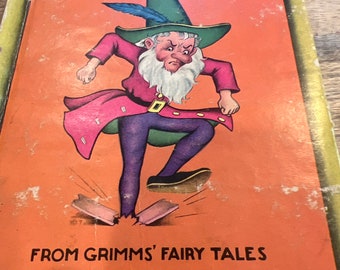 Puede incluir: Una portada de libro vintage con una ilustración de un gnomo de aspecto gruñón con un atuendo rojo y verde, un sombrero verde con una pluma y una barba blanca. El gnomo está pisoteando dos trozos de madera rotos. El título del libro es "Rumpelstiltskin" y el subtítulo es "From Grimms' Fairy Tales".