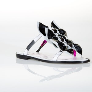 May include: White leather sandals with a black and white bow detail. The sandals have a flat sole and a pink accent.