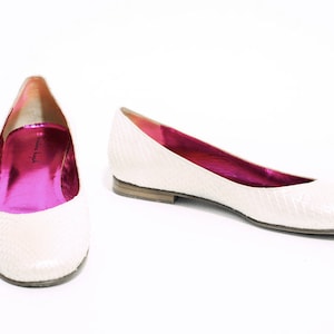 Könnte beinhalten: Ein Paar weiße Ballerinas mit Schlangenhautmuster und rosa Futter. Die Schuhe haben eine spitze Zehenpartie und einen niedrigen Absatz.