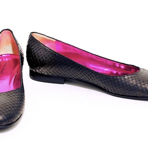 Pode incluir: Um par de sapatilhas de balé pretas de couro com padrão de pele de cobra. As sapatilhas têm um forro de cetim rosa.