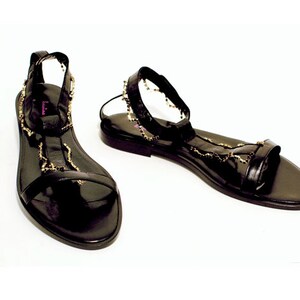 Puede incluir: Sandalias de cuero negro con detalles de cadena dorada. Las sandalias tienen una suela plana y una correa de tobillo.