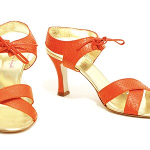 May include: A pair of orange leather heeled sandals with gold accents and a bow detail. The sandals have a criss-cross strap design and an open toe.